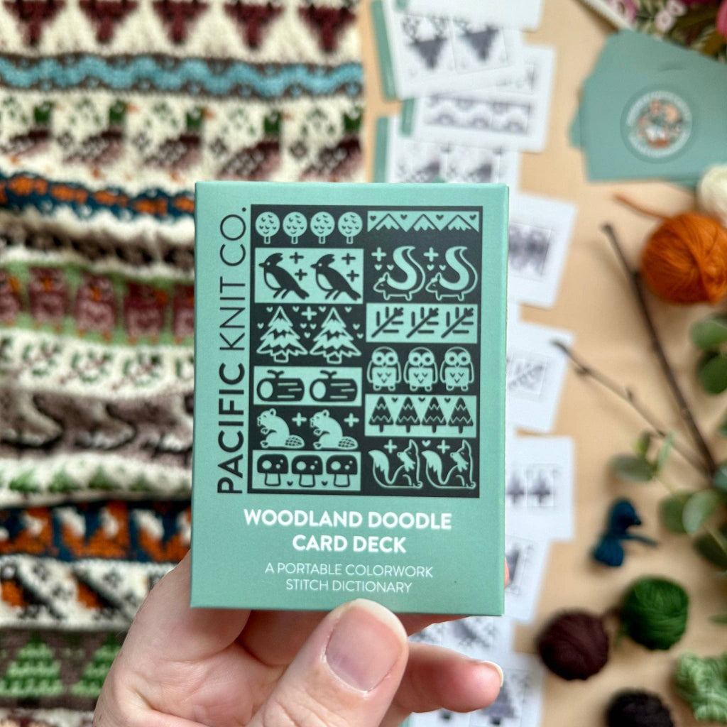 A hand holds a mint green box of the Pacific Knit Co. Woodland Doodle Card Deck, with a blurred background of knitting supplies and knitted patterns.