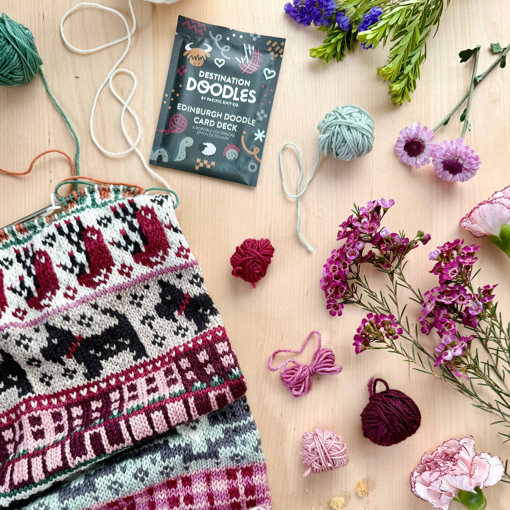 Knitted fabric with fair Isle pattern, yarn balls, flowers, and a card deck on a wooden surface.