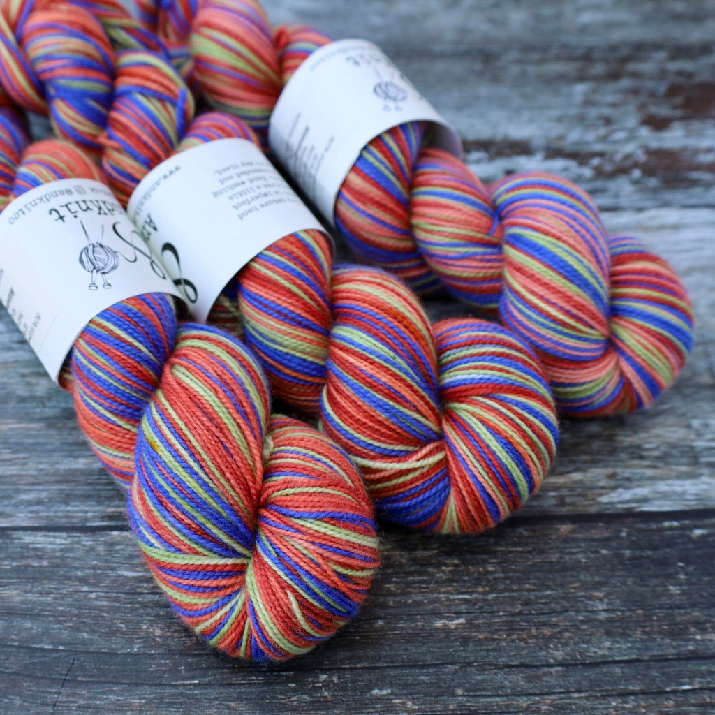 Several skeins of variegated yarn with white paper labels, featuring stripes of red, orange, blue, green, and yellow, arranged on a weathered gray wooden surface.