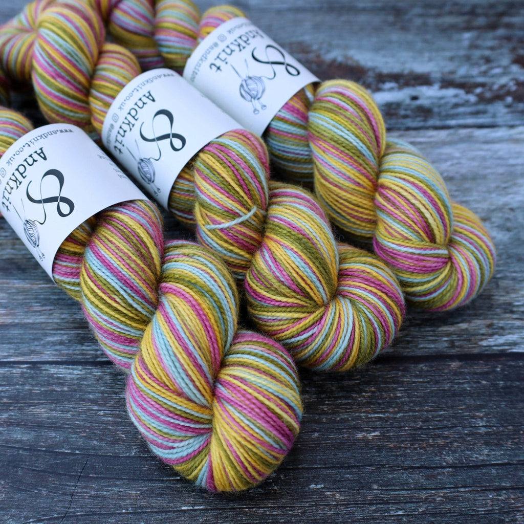 Three skeins of hand-dyed yarn with white labels reading AndKnit are arranged on a dark, weathered wooden surface.