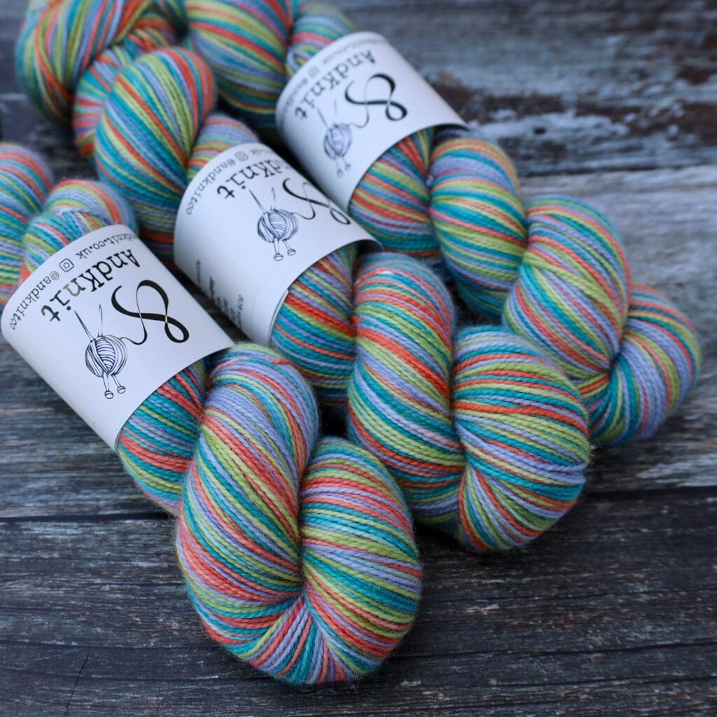 Three skeins of knitting yarn with white paper bands, featuring a variegated multicolour pattern of teal, green, blue, orange, pink, and yellow, arranged on a dark rustic wooden surface.