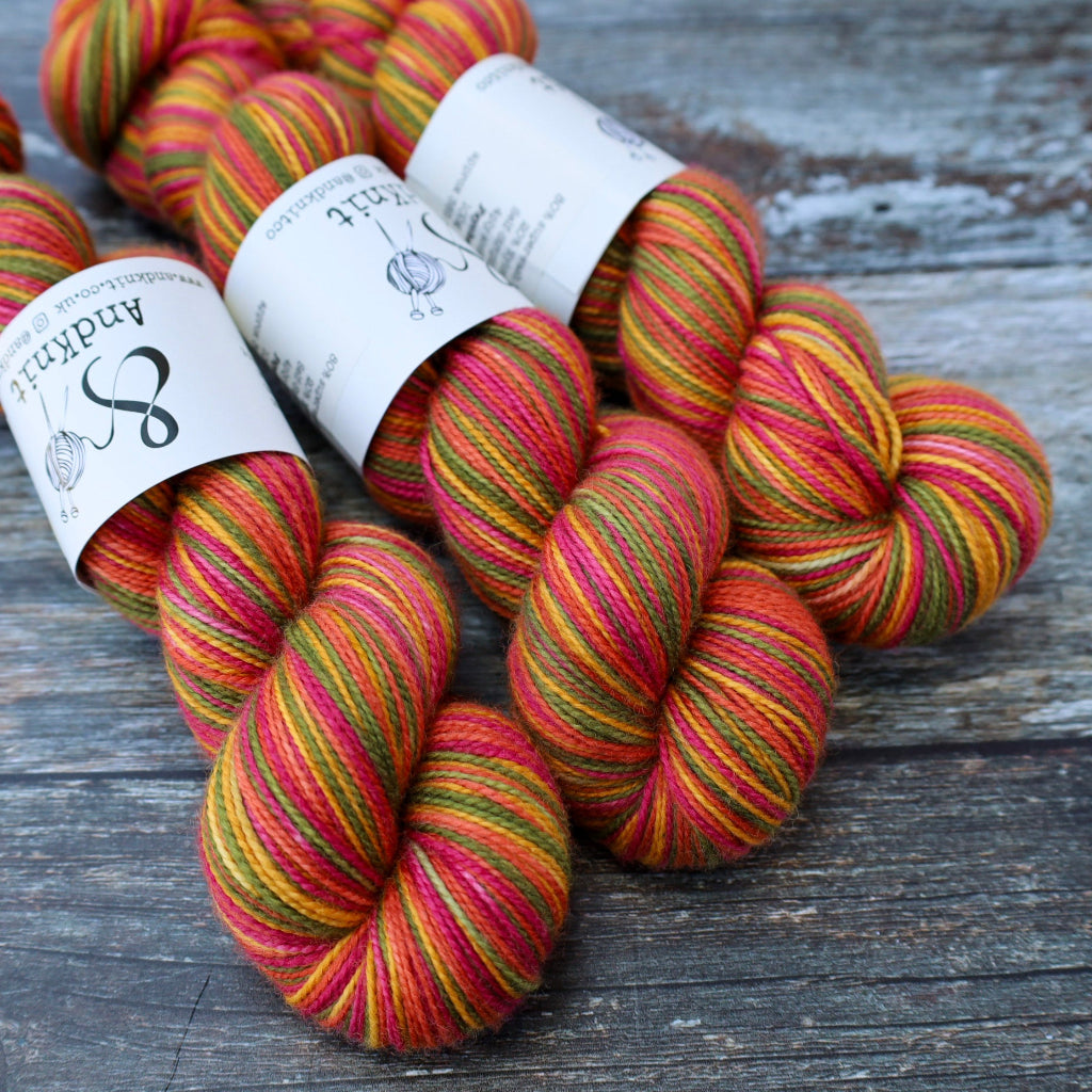 Three skeins of AndKnit yarn with white paper labels, featuring variegated colours of red, pink, orange, yellow, and green, arranged on a rustic dark grey wooden surface.