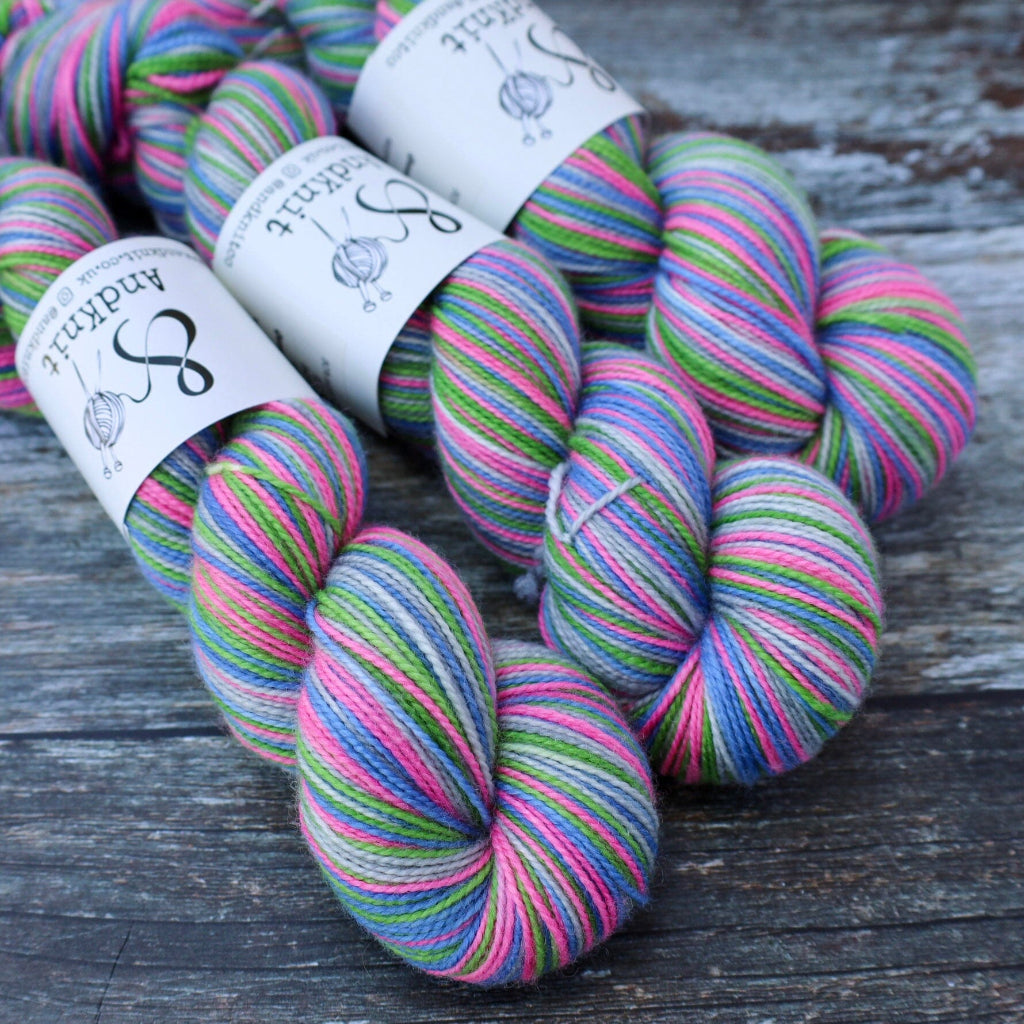 Several skeins of Andknit variegated yarn with white paper labels, displayed on a rustic dark wood surface. The yarn features a vibrant, multi-coloured variegated pattern of pink, green, blue, and purple.