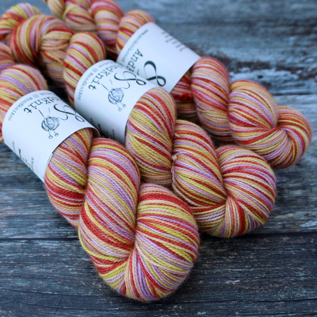 Three skeins of variegated yarn with white paper bands, featuring stripes of red, orange, yellow, pink, and cream, arranged on a weathered gray wooden surface.