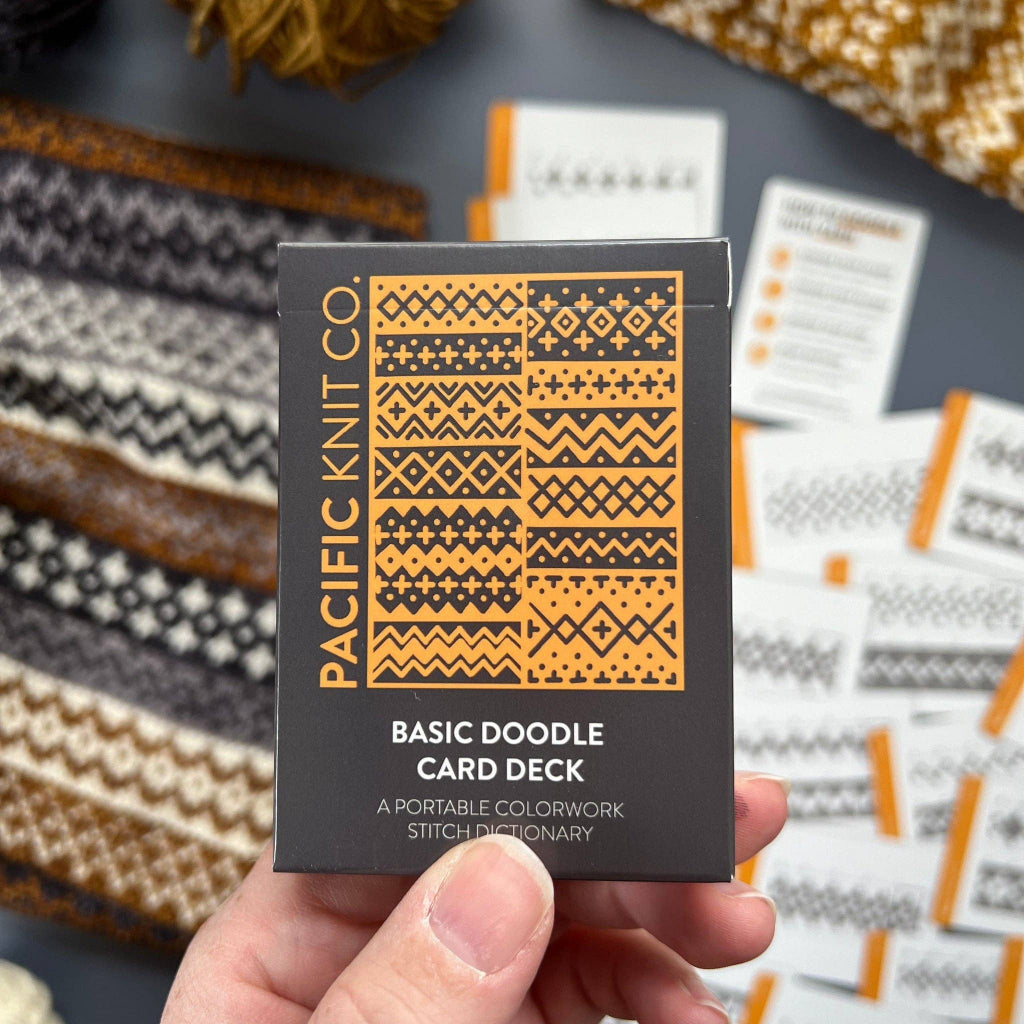 A hand holds a black card deck box labeled 'PACIFIC KNIT CO. BASIC DOODLE CARD DECK' with orange geometric patterns on the front.