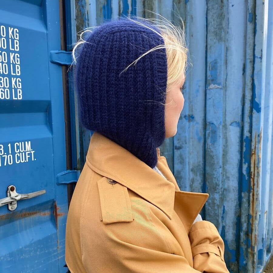 A person wearing a dark navy blue chunky-knit balaclava hood with ear flaps, paired with a tan trench coat, standing in front of a weathered blue shipping container.