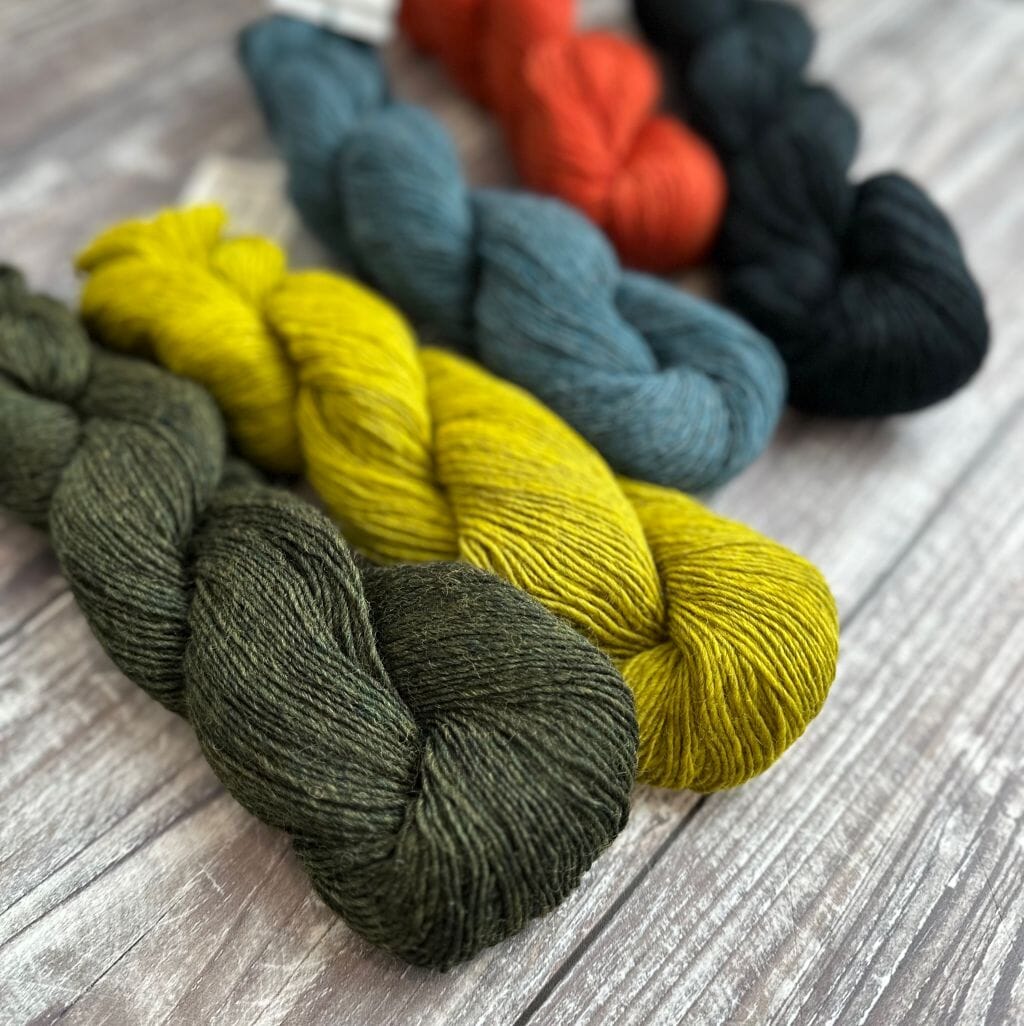 Several skeins of Blue Sky Fibers Woolstok Light yarn in various colors including yellow, green, blue, and black, displayed on a wooden surface.