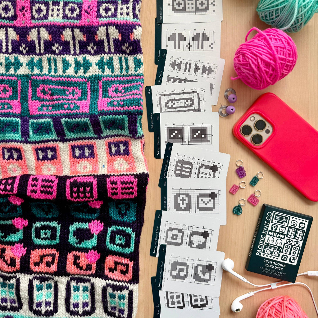 A styled flat-lay of knitting supplies on a wooden surface, featuring a colorful knitted scarf with geometric patterns, a stack of white pattern cards with pixel-art style icons, balls of pink, teal, and purple yarn, a red smartphone case, white earbuds, and a small black card with a QR code.