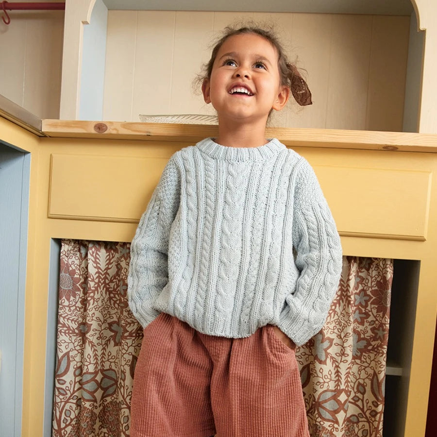 Child wearing a light blue sweater and pink shorts standing in front of a wooden cabinet with patterned curtains.
