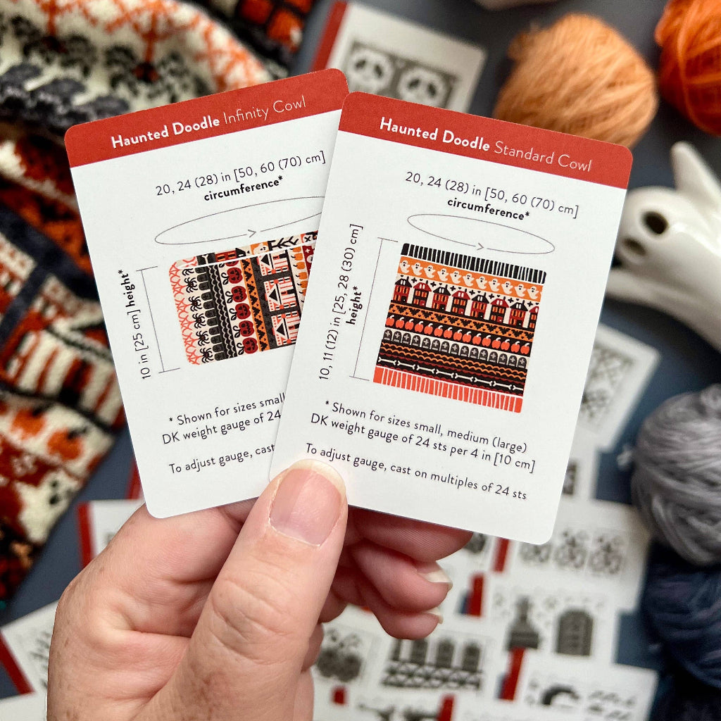 A hand holds two knitting pattern cards for 'Haunted Doodle' yarns, with a cozy, knitted blanket and yarn balls blurred in the background.