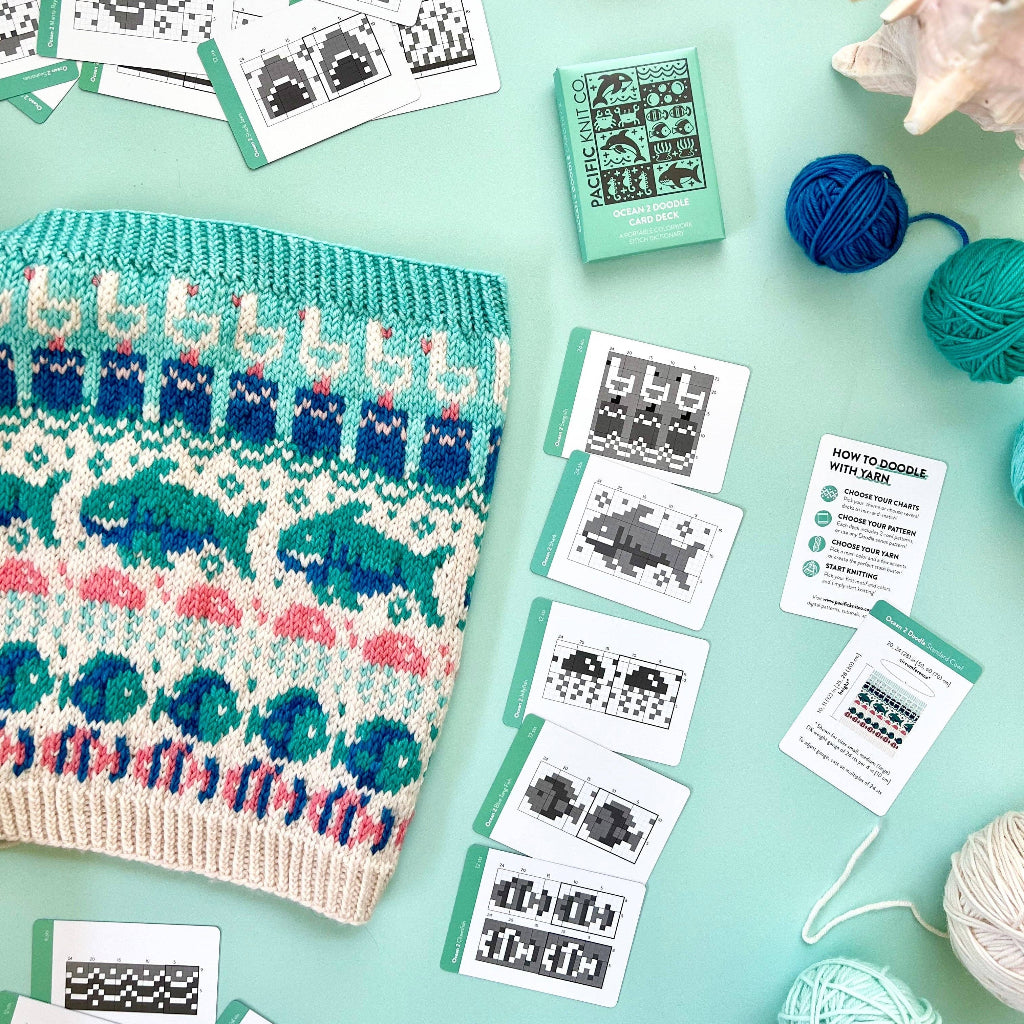 A flat-lay of a knitted sweater with a colorful Fair Isle pattern, surrounded by knitting cards, balls of yarn, and a small mint green book on a light aqua background.