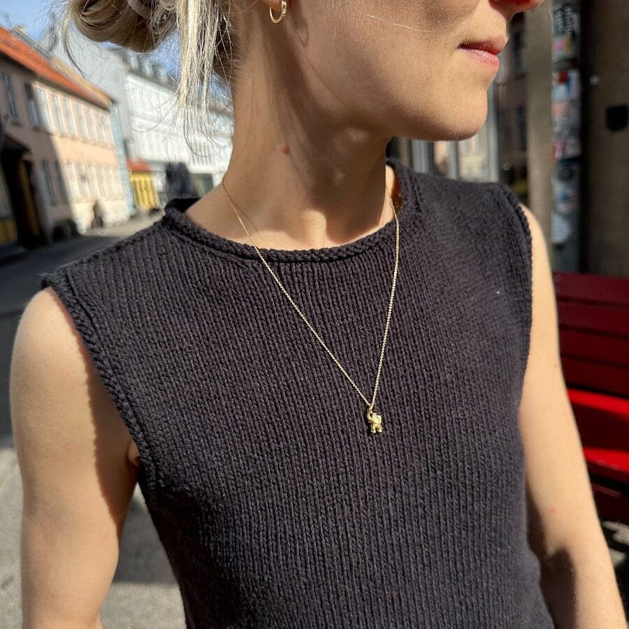 A close-up lifestyle shot of a woman wearing a dark charcoal ribbed knit sleeveless top and a delicate gold necklace with a small animal charm, set against an outdoor street background.