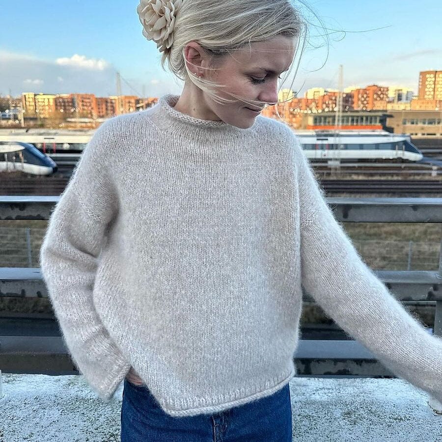 Woman wearing a light sweater and blue jeans standing on a rooftop with a cityscape in the background.