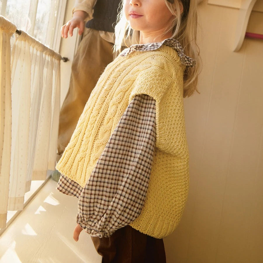 Child wearing a yellow knitted vest and checkered shirt standing indoors.