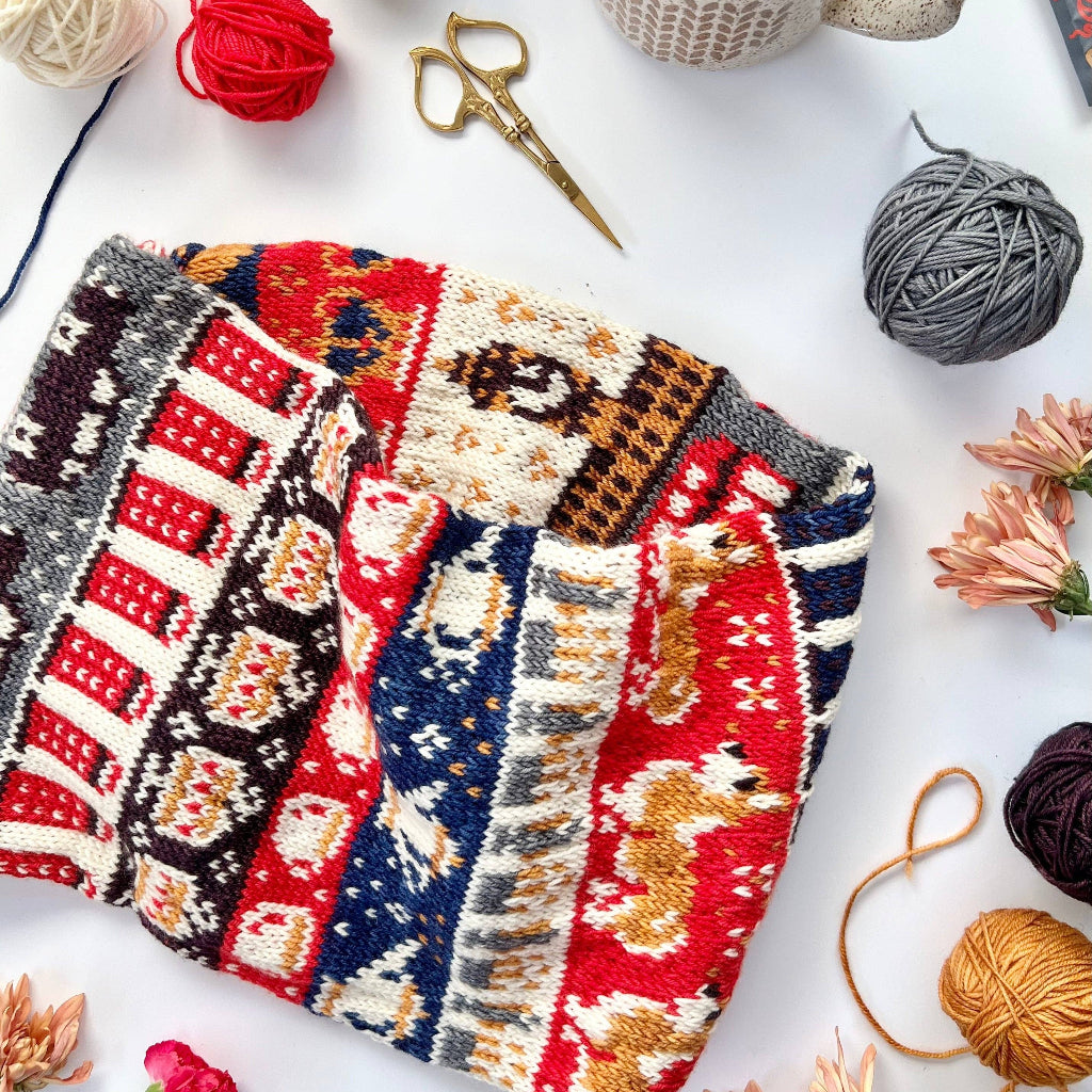 Knitted cowl with London Doodle Designs surrounded by yarn balls, and flowers on a white surface.