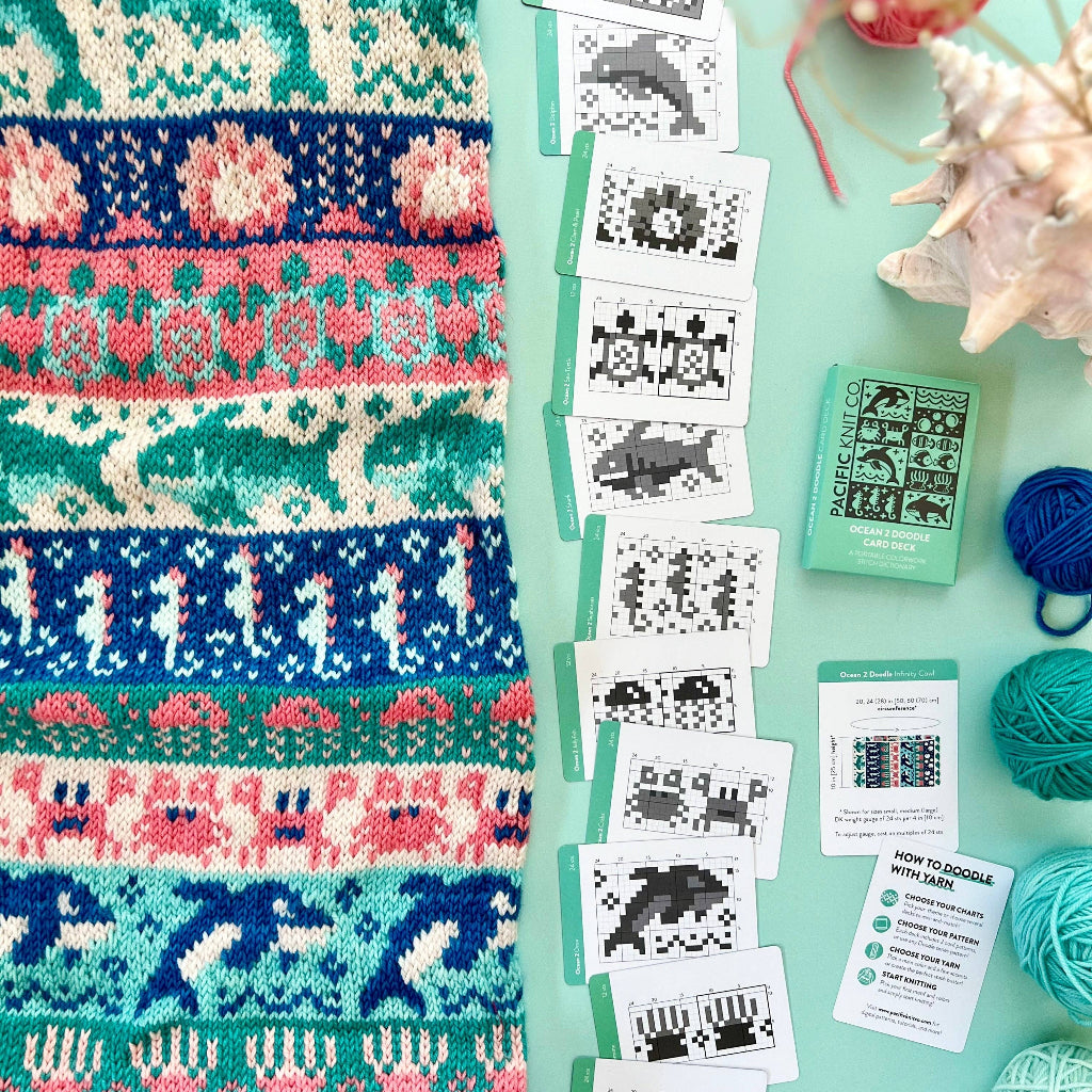 A hand holds a mint green box labeled 'PACIFIC KNIT CO. OCEAN 2 DOODLE CARD DECK' against a styled flat-lay background with colorful yarn balls, fabric, and sewing supplies.