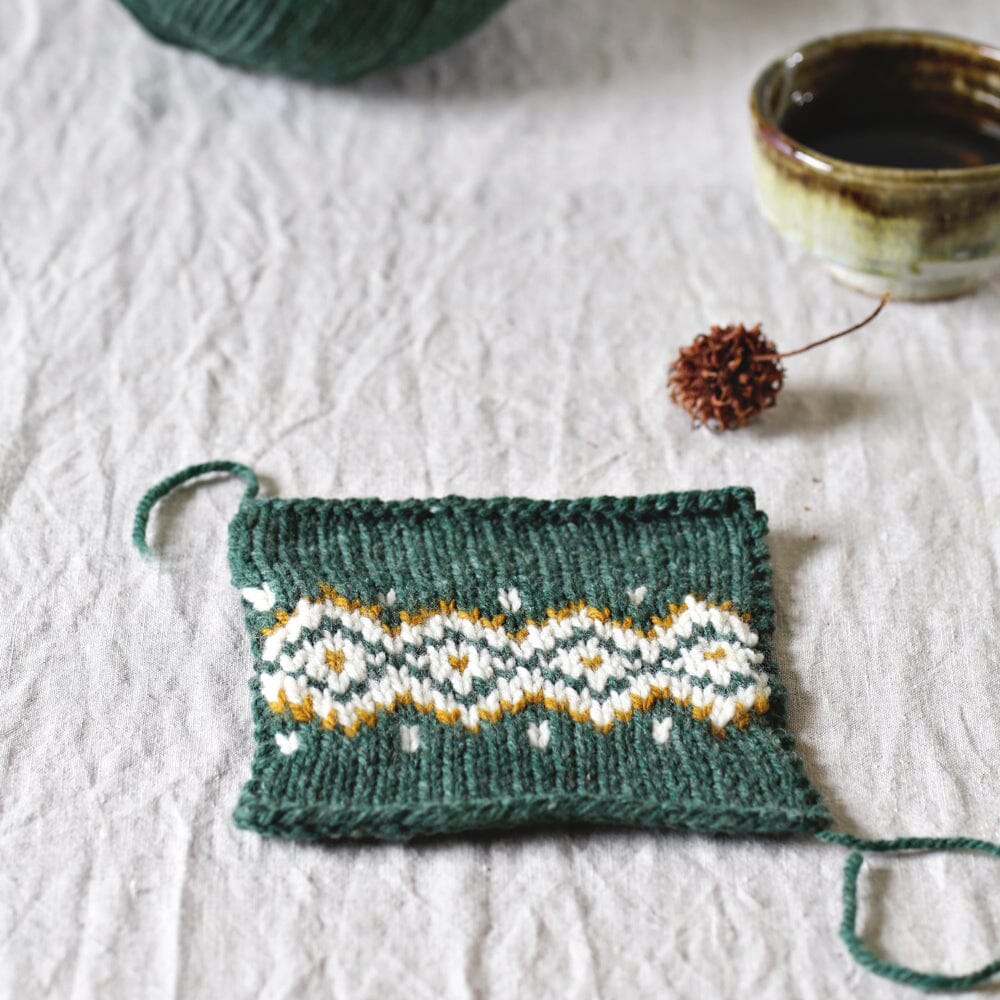 Knitted swatch with a pattern on a textured surface with yarn and a bowl.