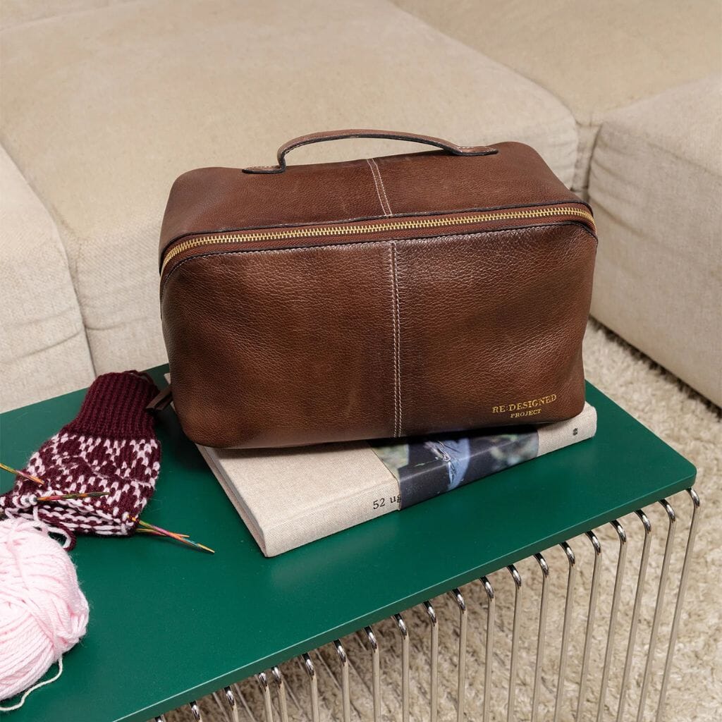 Brown leather project bag  on a green table with books and knitting items.