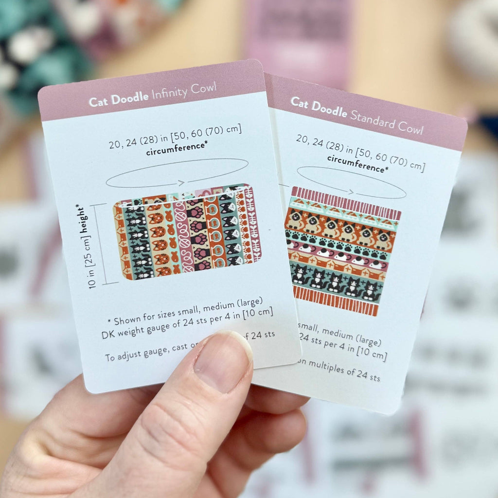 A hand holds two printed knitting gauge cards in front of a blurred craft supply background. The cards are labeled 'Cat Doodle Infinity Cowl' and 'Cat Doodle Standard Cowl' with knitting pattern diagrams and measurements.