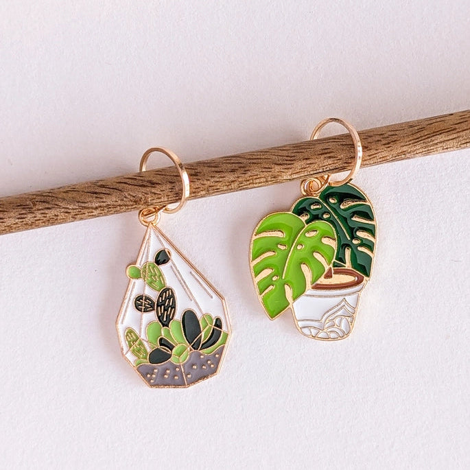 Two gold-tone enamel plant-themed earrings hanging from a wooden stick against a white background. One earring features a cactus in a geometric pot, and the other features green monstera leaves in a potted style.