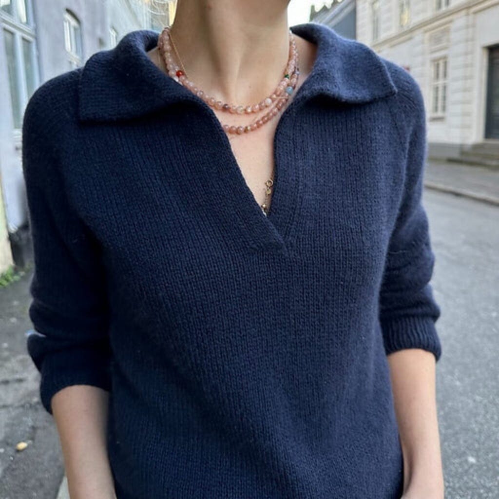 A woman wearing a dark navy blue knit polo sweater, layered with a peach-colored beaded necklace and gold jewelry, standing outdoors on a city street.