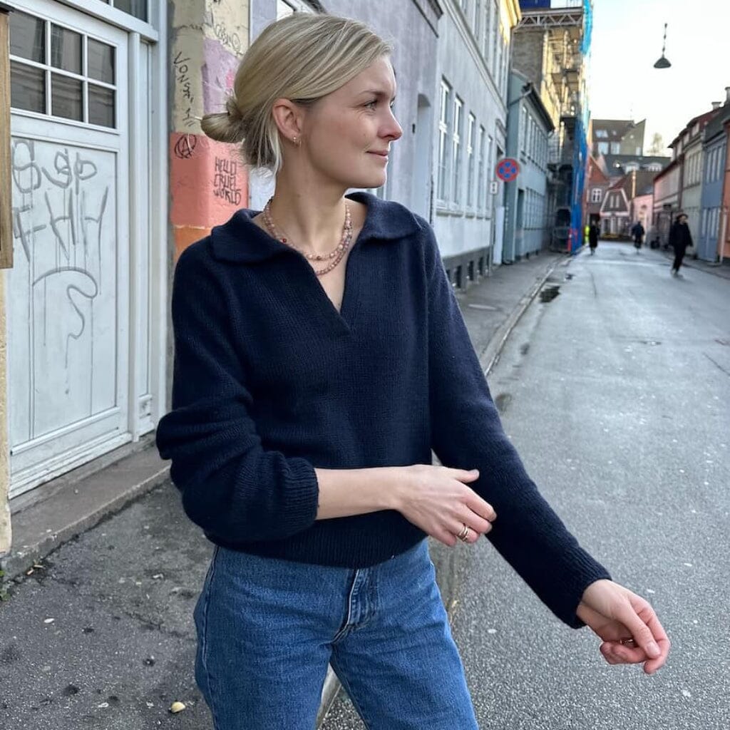 A woman with blonde hair tied back wears a dark navy blue knit zip-up hoodie and blue jeans, standing on a city street.