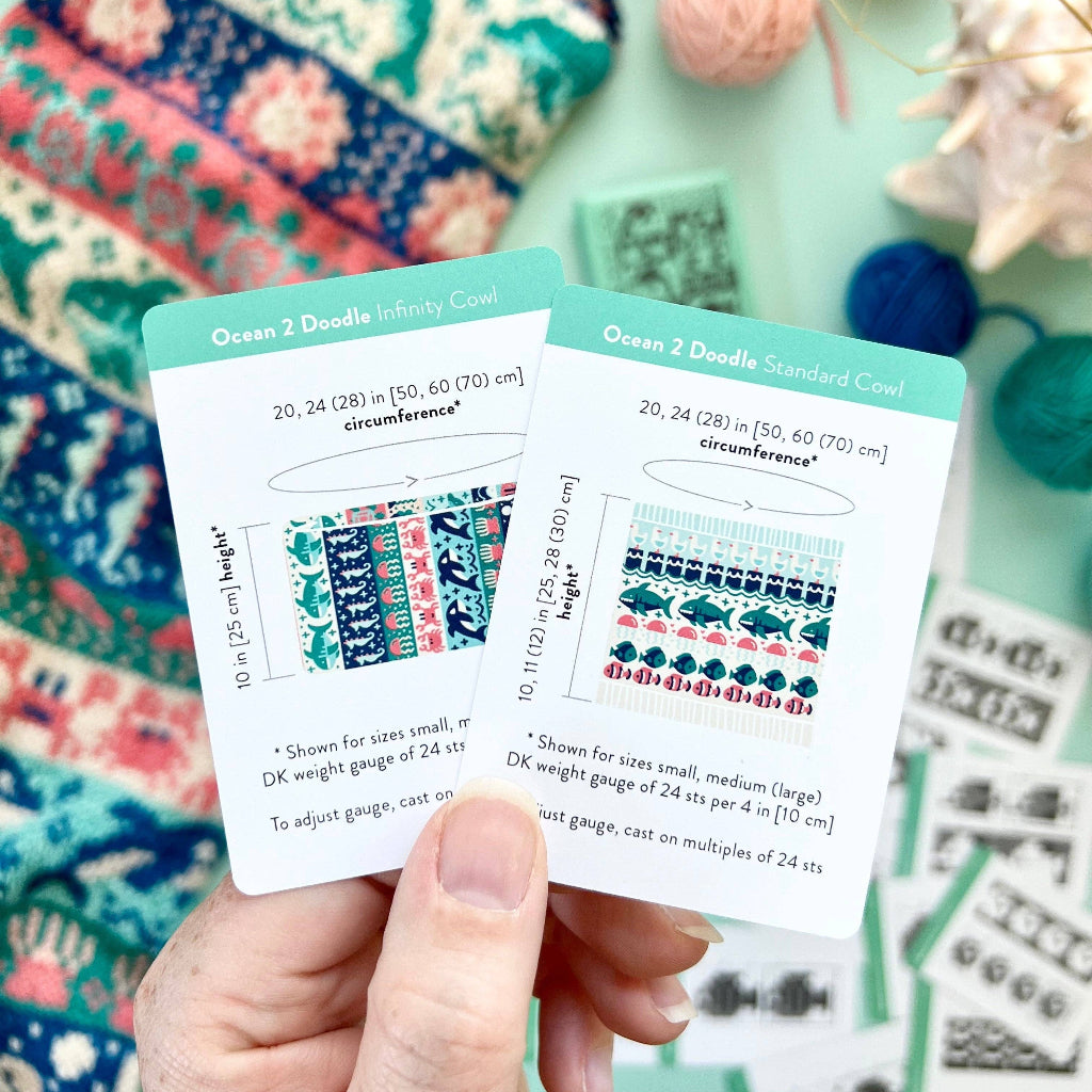 A hand holds two knitting pattern cards for 'Ocean 2 Ddouble Infinity Co' and 'Ocean 2 Ddouble Standard Cowl' against a styled background of colorful knitted fabrics, yarn balls, and knitting needles.
