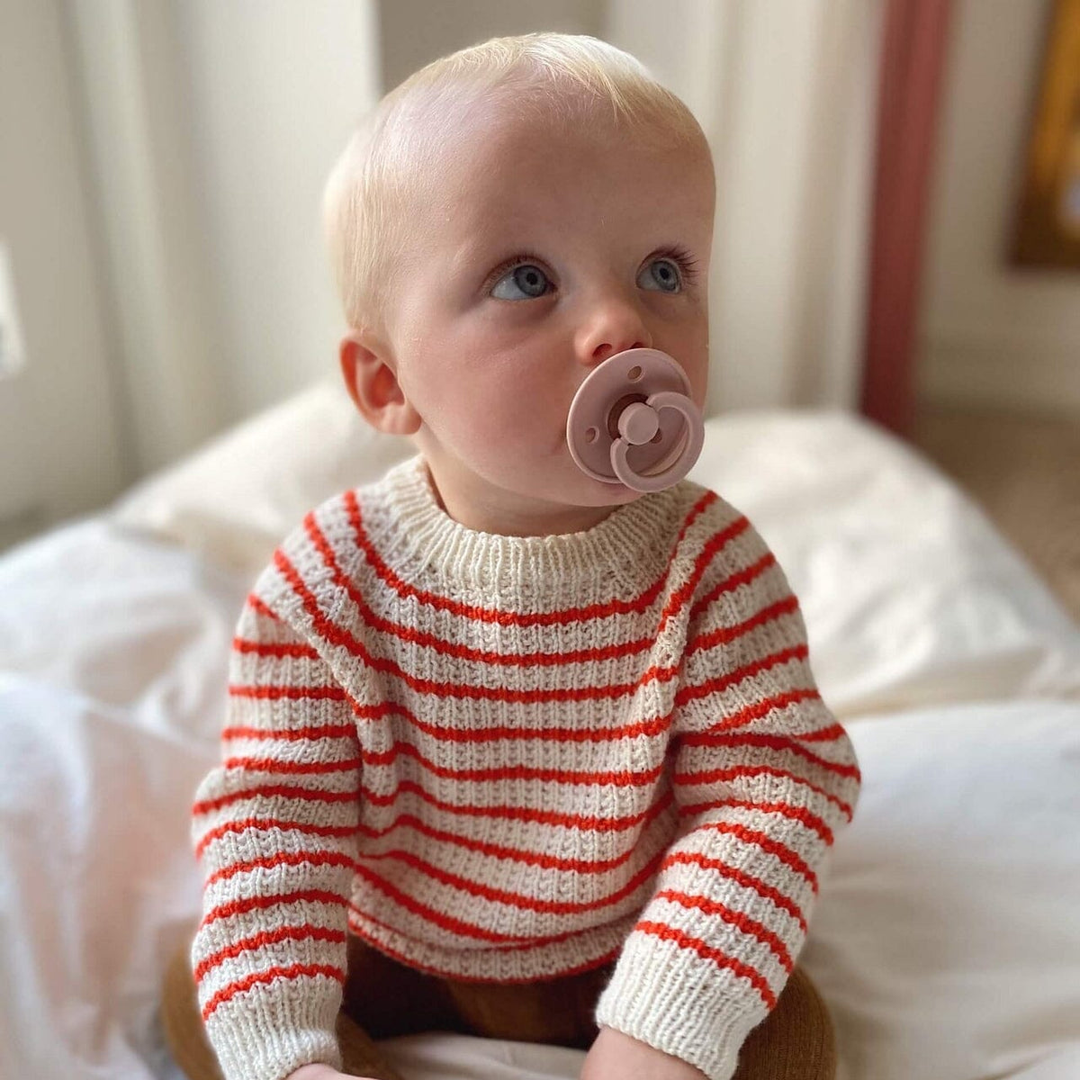 Baby wearing a red and white striped sweater with a pacifier, sitting on a bed.