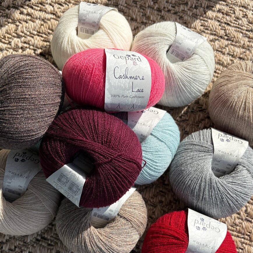 A group of Gepard Cashmere Lace yarn skeins in various colors, including cream, brown, pink, burgundy, light blue, gray, and red, arranged on a textured woven rug.