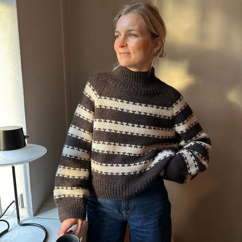 A woman wearing a dark brown and cream striped knit sweater with blue jeans, standing in a sunlit room next to a small side table.