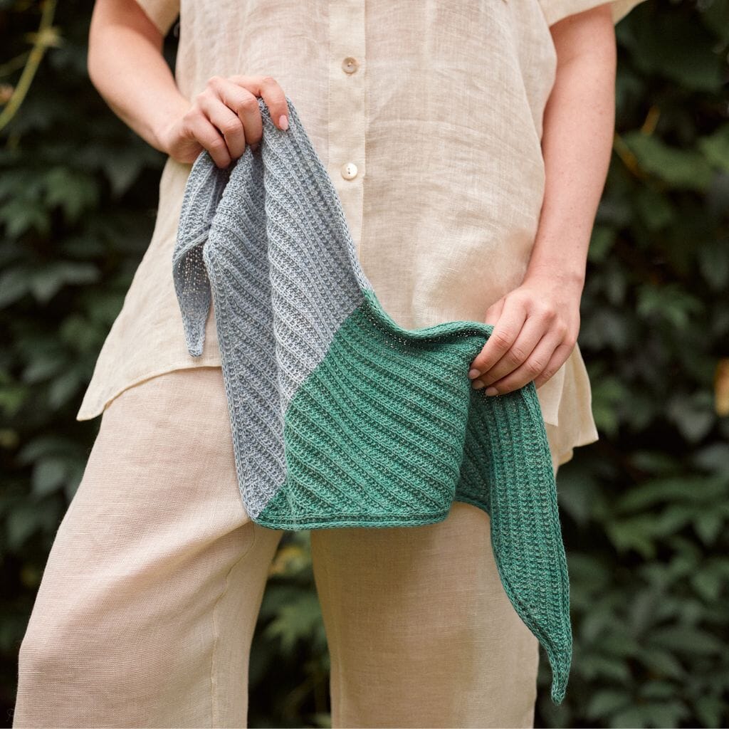 Person holding a green and gray knitted shawl against a blurred natural background