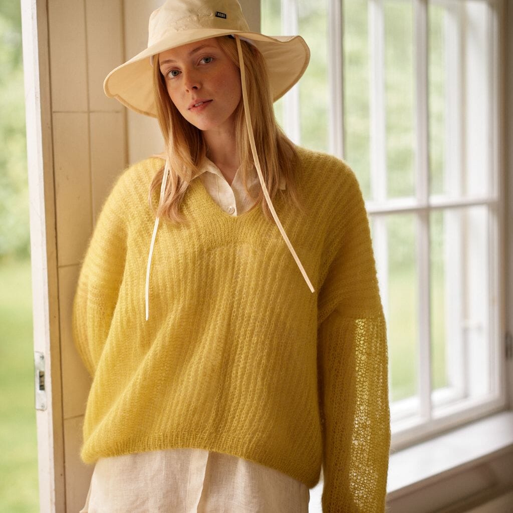 Woman wearing a yellow sweater and beige hat indoors near a window