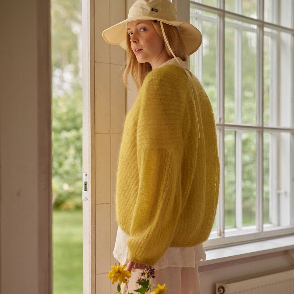 Woman wearing a yellow sweater and beige hat standing by a window with a garden view.