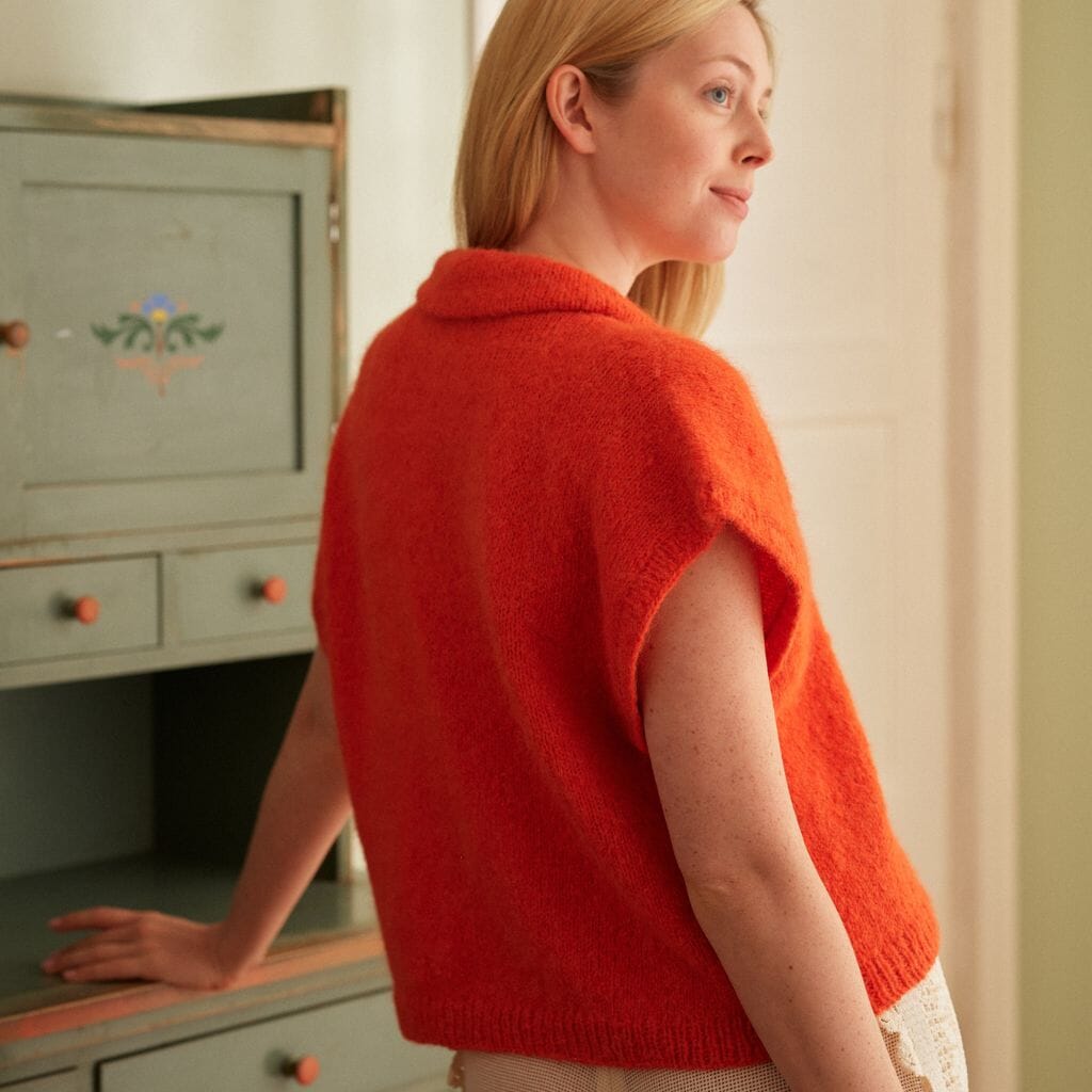 Woman wearing a red knitted vest standing in a room with a cabinet.