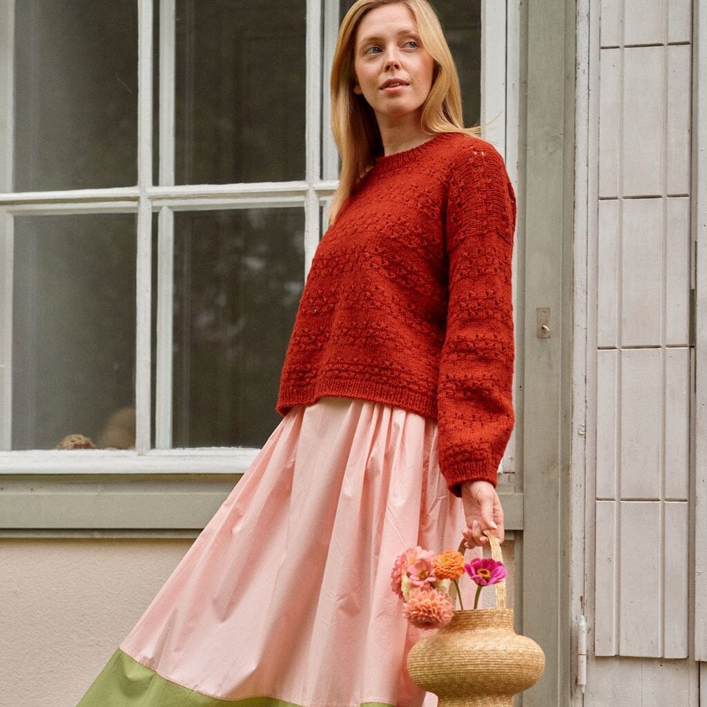 Woman wearing a red sweater and pink skirt holding a basket with flowers, standing outside a house.