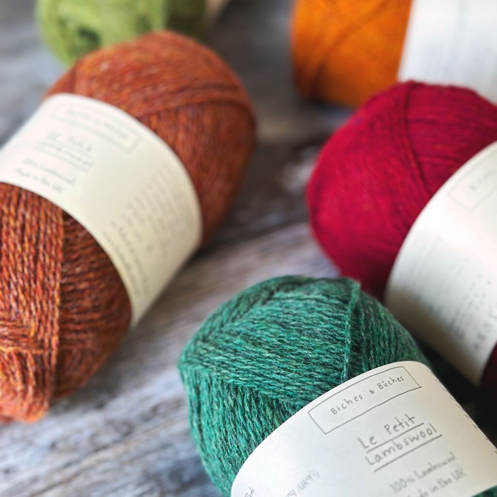 Several skeins of Biches &amp; Büches Le Petit Lambswool yarn in various colors arranged on a wooden surface.