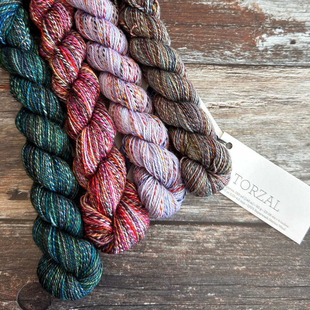Four skeins of variegated yarn arranged on a rustic wooden surface, with a white paper tag attached. The yarn colors include teal, pink, red, purple, and brown, and the tag shows the brand name TORZAL.