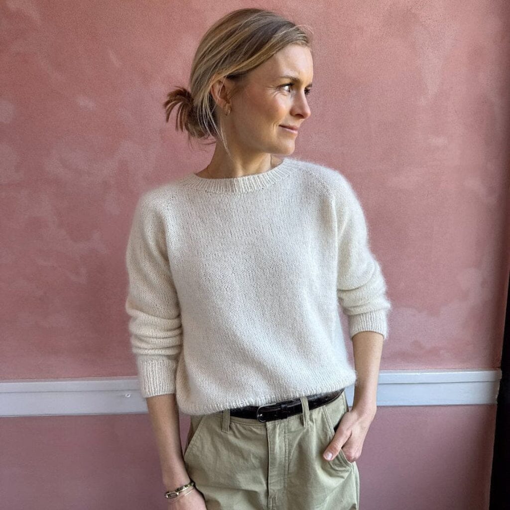Woman wearing a light-colored sweater and beige pants against a pink wall.