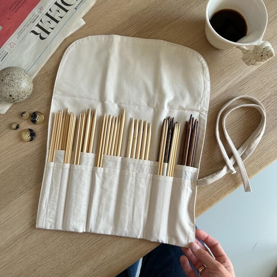 Set of wooden knitting needles in a white case on a wooden table with a newspaper and cup of coffee.