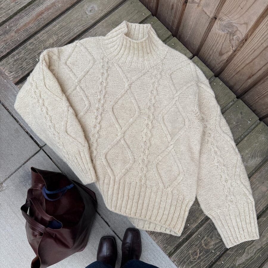 White cable knit sweater on a wooden deck with a brown bag and dark shoes.