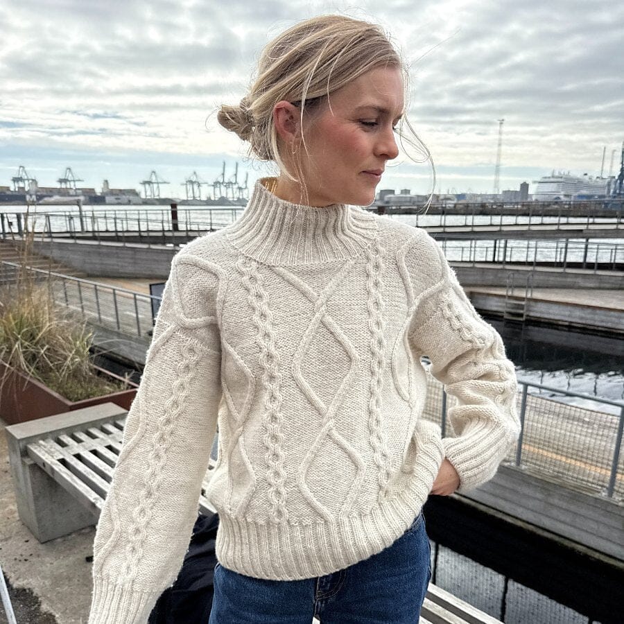 Woman wearing a cream cable knit sweater and blue jeans on a rooftop with a cityscape background.