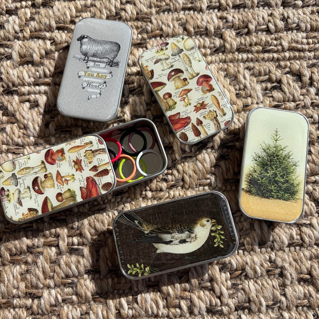 Five small metal tins with illustrated vintage-style covers are arranged on a woven brown rug. One tin is open, showing several round ring-shaped items inside, and the others display designs including a sheep, mushrooms, fish and bird motifs, and a pine tree.