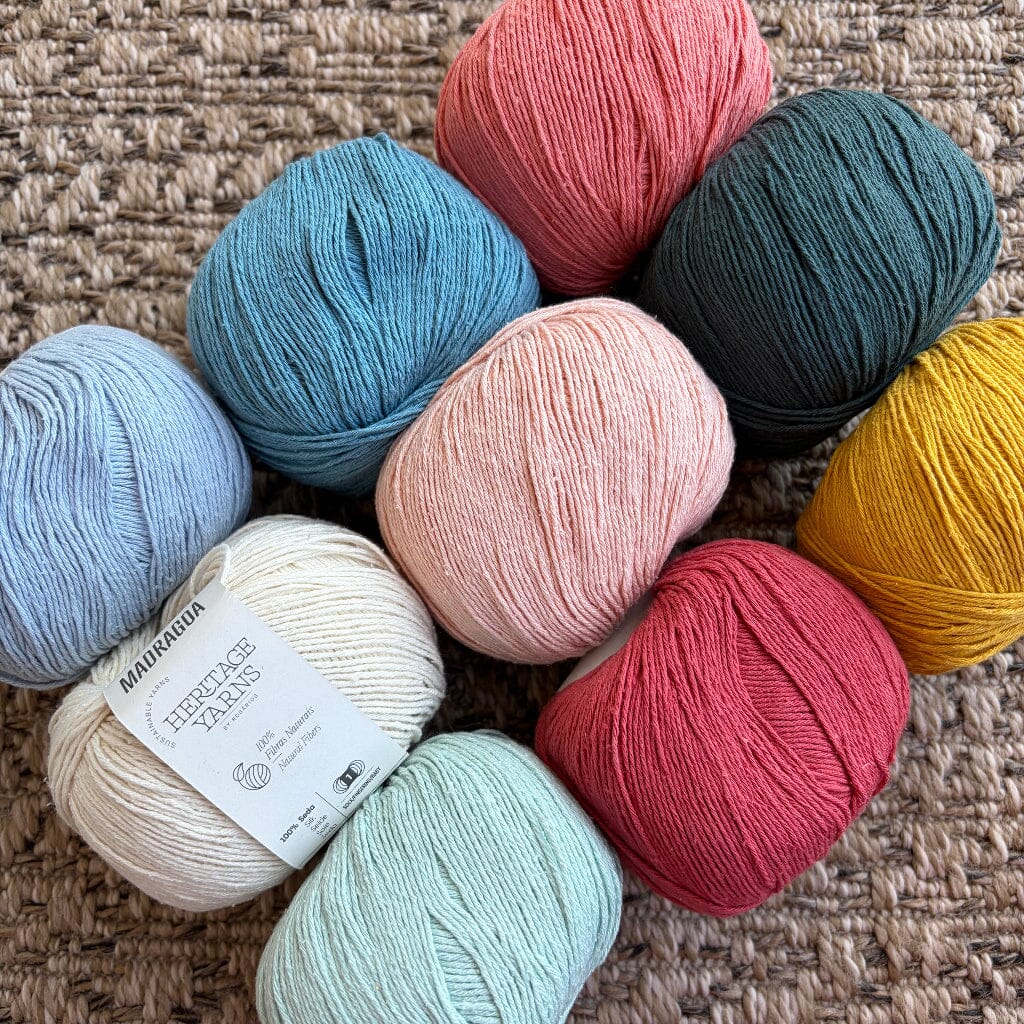nine  balls of textured yarn in assorted pastel and earthy colors, including mustard yellow, teal, dusty pink, blush, pale pink, dusty blue, mint green, cream, and light blue, arranged on a woven beige rug. A white paper label is attached to one of the lower-center balls.
