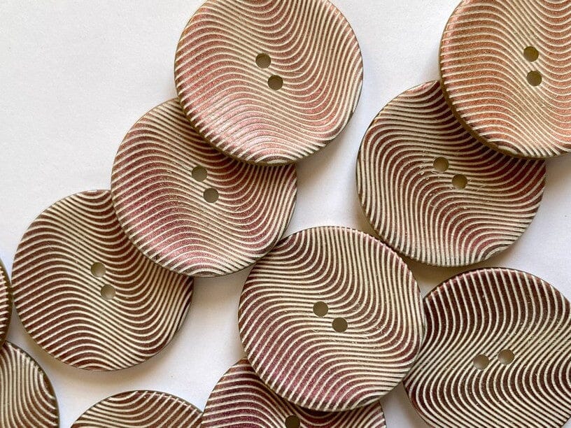 A close-up of several round wooden buttons with two holes, featuring a carved wavy pattern in alternating light beige and reddish-brown tones, arranged on a plain white background.