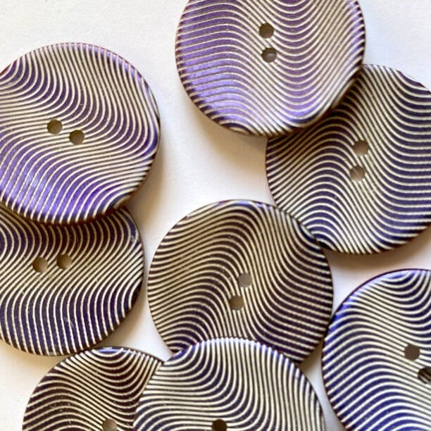 A close-up of multiple round decorative buttons with two holes, featuring a wavy striped pattern in black, white, and a soft purple/lavender tone, arranged on a plain light background.