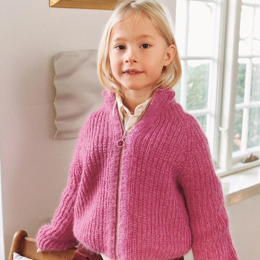 Child wearing a pink knitted cardigan sitting on a chair indoors.