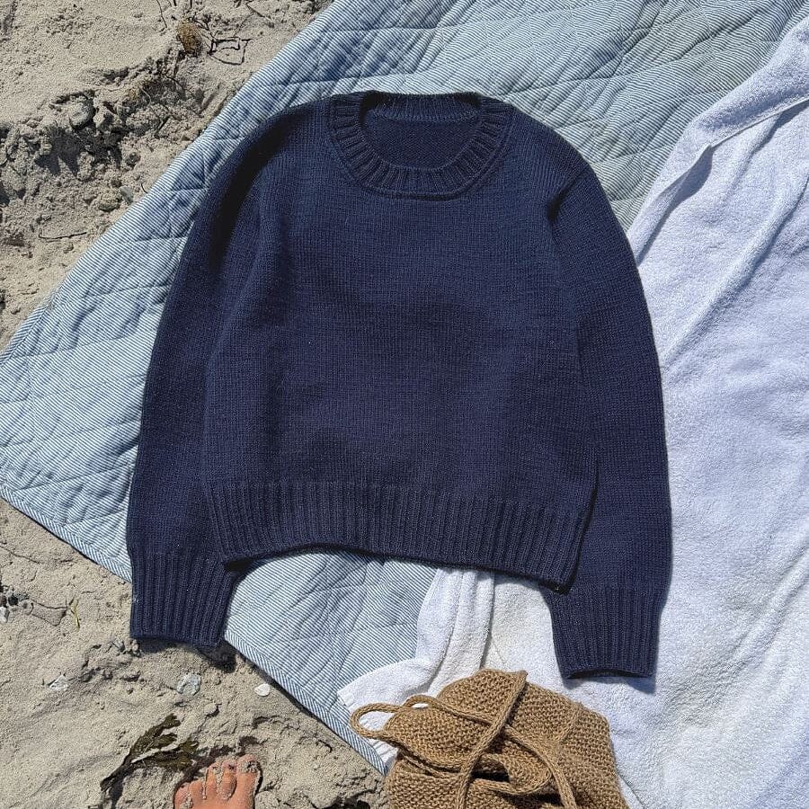 A navy blue knit crewneck sweater laid flat on a beach towel on sand, with bare feet visible at the bottom left and a small woven bag nearby.