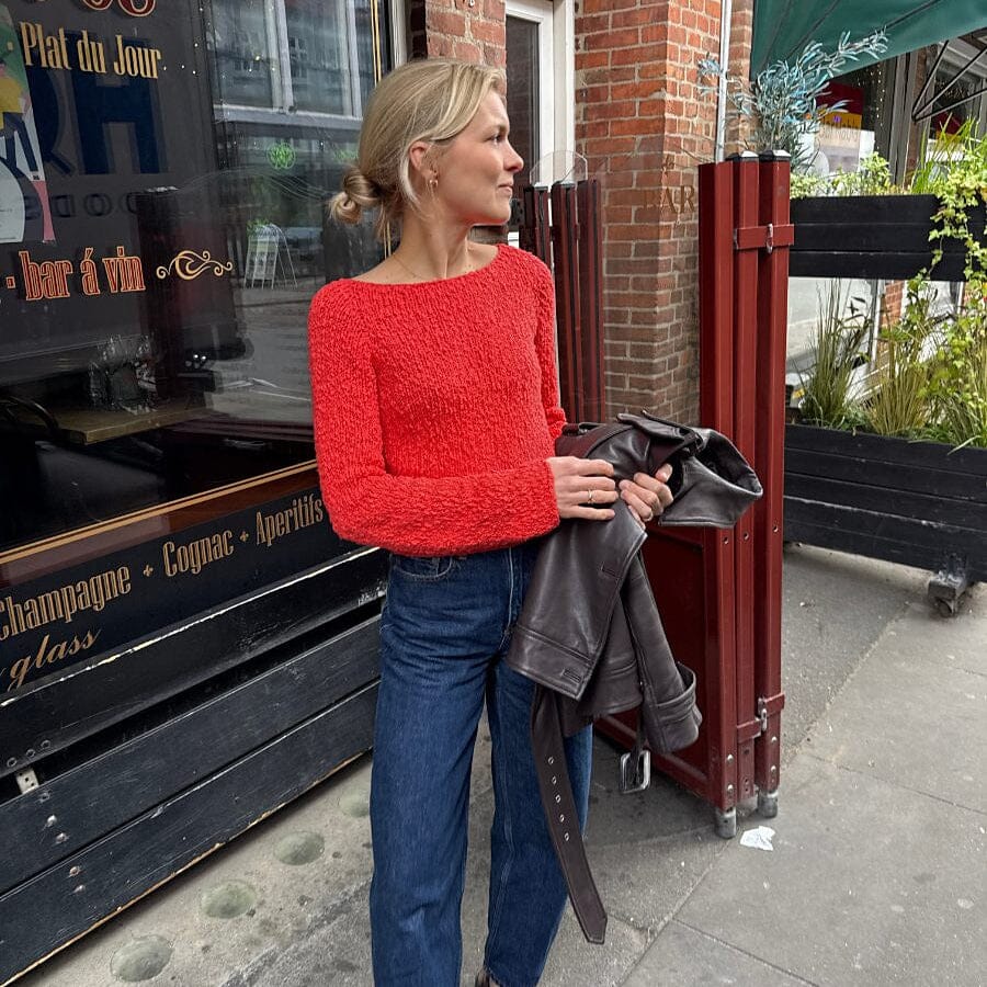 Woman in a red sweater and blue jeans holding a gray jacket on a street.